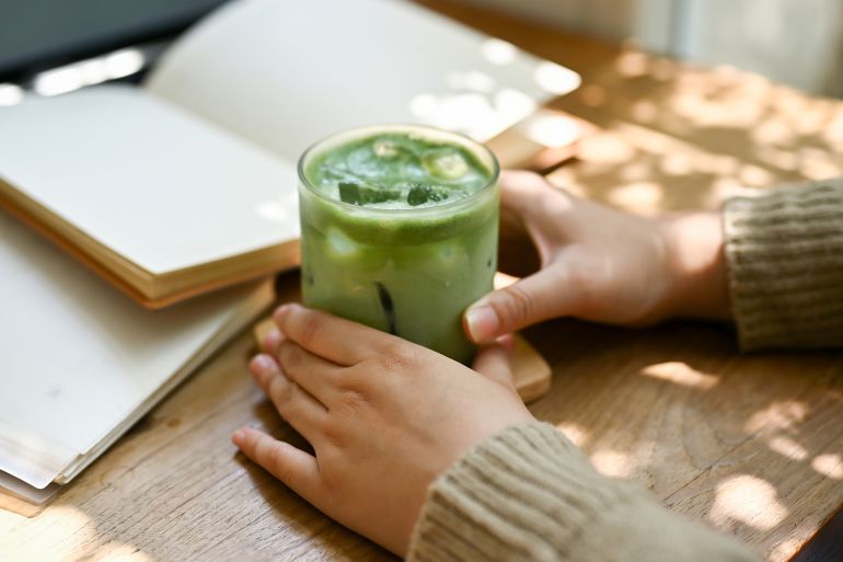 Close-up image of a female in comfy sweater holding a glass of iced matcha green tea at a table in a beautiful minimal cafe.; Shutterstock ID 2298328383; purchase_order: aj; job: ; client: ; other: