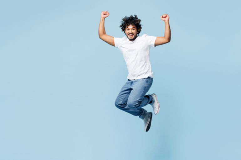 Full body overjoyed young fun happy Indian man he wear white t-shirt casual clothes jump high do winner gesture isolated on plain pastel light blue cyan background studio portrait. Lifestyle concept