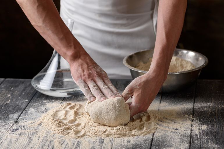 The process of preparing dough. A cook kneads dough from whole grain wheat flour in the kitchen. Water and flour on a wooden table. Baking baked goods or pizza. Chef's hands close-up.