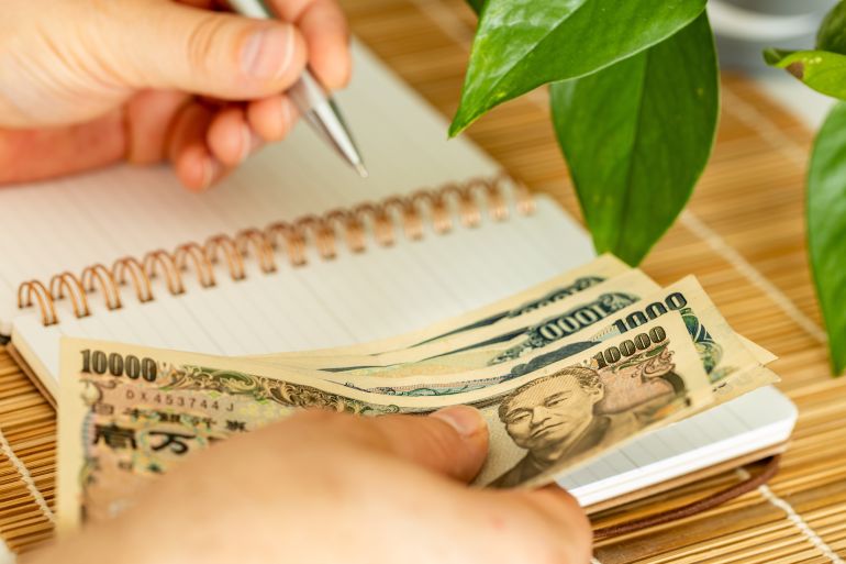 A Japanese man holding money in his hand and writing down his income and expenses in a notebook. A Japanese way of saving and controlling a household budget called kakeibo. Japan finances. copy space
