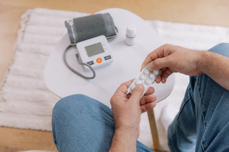 Close-up of elderly man sitting sofa at living room measures blood pressure with device. Senior male holding pills and drops drugs. Health and self-care concept, copy space