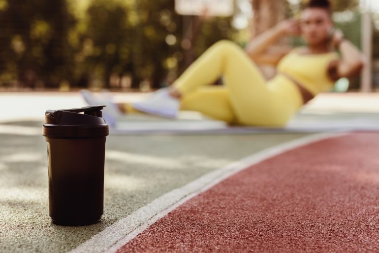 Black woman doing abs exercises crunches with arms behind head on floor yoga mat, working on six-pack abs in blurred background, selective focus on black shaker on basketball field ground