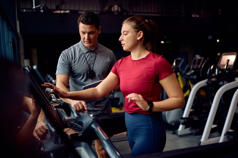 Personal trainer monitoring the running of athletic woman on treadmill during sports training in a gym.