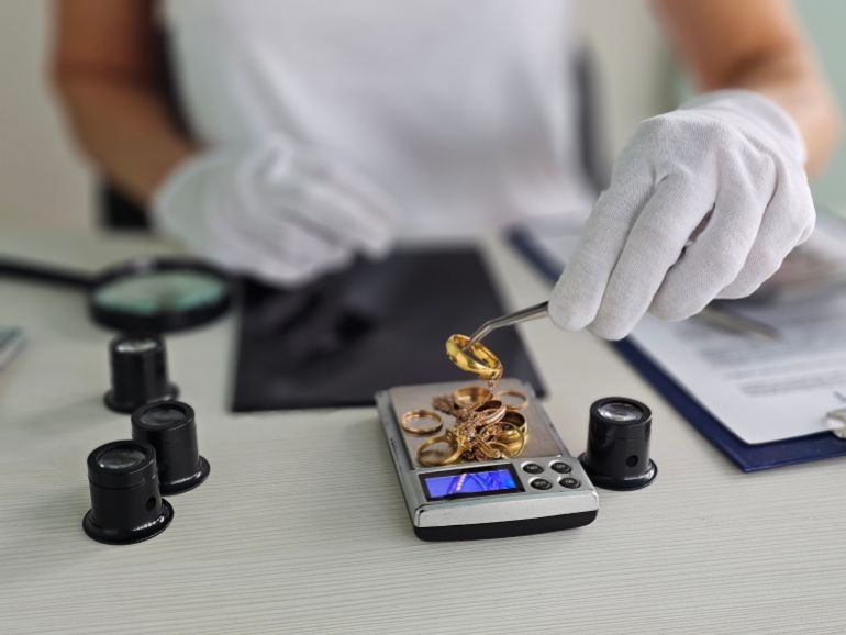 A person wearing gloves measures various gold jewelry pieces on a digital scale in an office