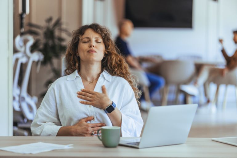 A professional woman seated at her desk practices mindfulness and relaxation, promoting workplace wellness. The scene emphasizes the importance of stress management and mental health in a busy office