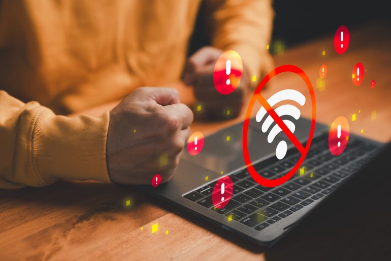 shutterstock_2681570265-1772288153 Frustrated user with laptop facing WiFi disconnection, red warning icons and no internet signal error symbol. Concept of network outage, connectivity problem, unstable internet and online frustration.
