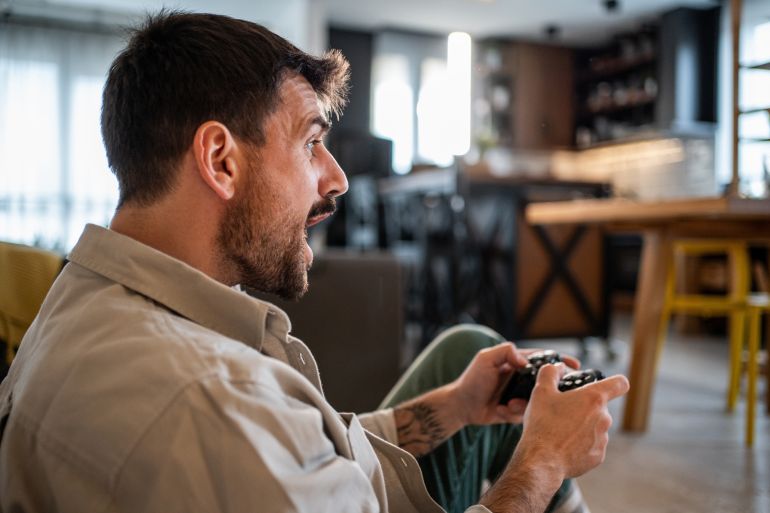 Young man engrossed in playing video games, holding a wireless controller with intense focus and an open mouth expression, enjoying his leisure hobby at home