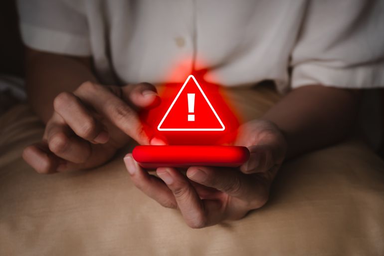Hacker attack detection alert, sms fraud and scammer calls, fraudulent phone calls concept. Red triangle warning caution alert show on smart mobile phone in senior elderly hands, in the dark room.
