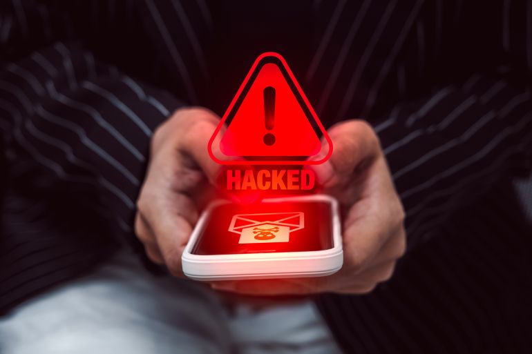 8967-1776418186 Close-up of white smartphone in person hand with hacked warning symbol and virus icon, showing phone scam, email or SMS phishing attack, mobile malware, cybersecurity risk, and digital data fraud.