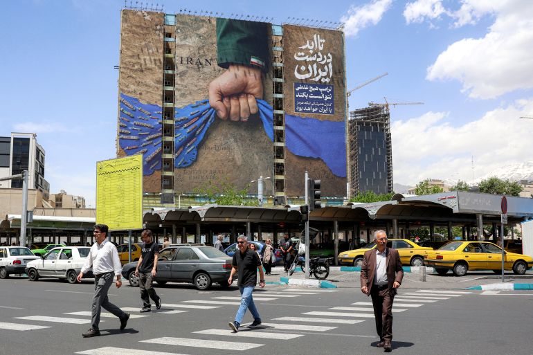 People cross a street near a billboard on the facade of a building depicting the Strait of Hormuz with a caption in Persian reading "Forever in Iran’s Hand", at Vanak Square in Tehran on May 6, 2026.
