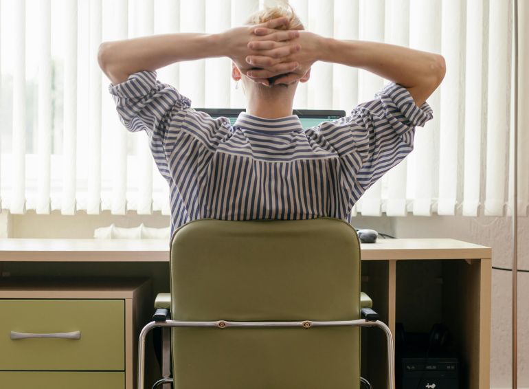A person sitting at a desk in an office, viewed from behind with hands behind head.