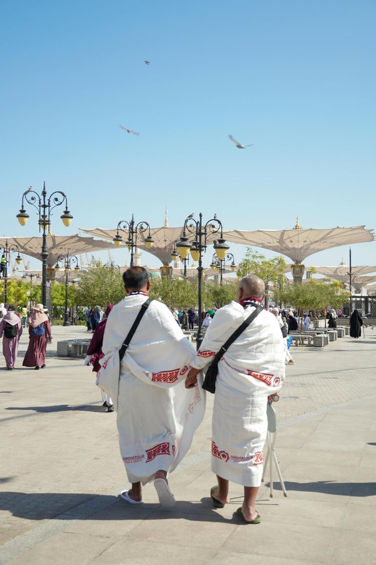 alhj-1778757146 Pilgrims in traditional attire walking outdoors in Medina, Saudi Arabia with umbrellas in the background.