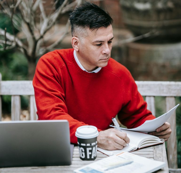 Adult man in red sweater working remotely at outdoor cafe table with laptop and coffee.