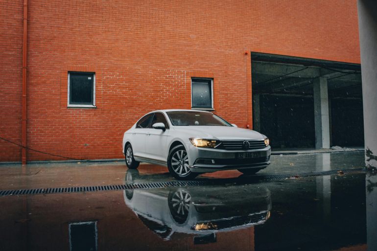 basat-1778939169 White sedan reflected on wet pavement against a red brick wall, showcasing modern automotive design.