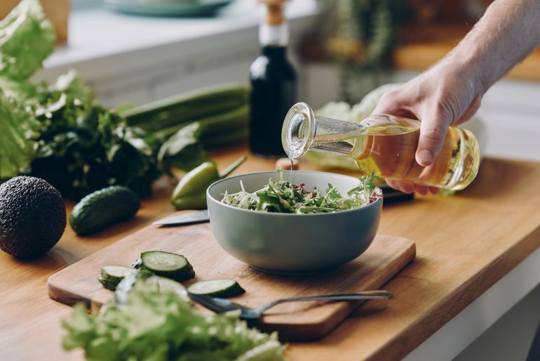 gettyimages-1429975622-1773543818 Close-up of unrecognizable man pouring olive oil into the bowl with fresh salad