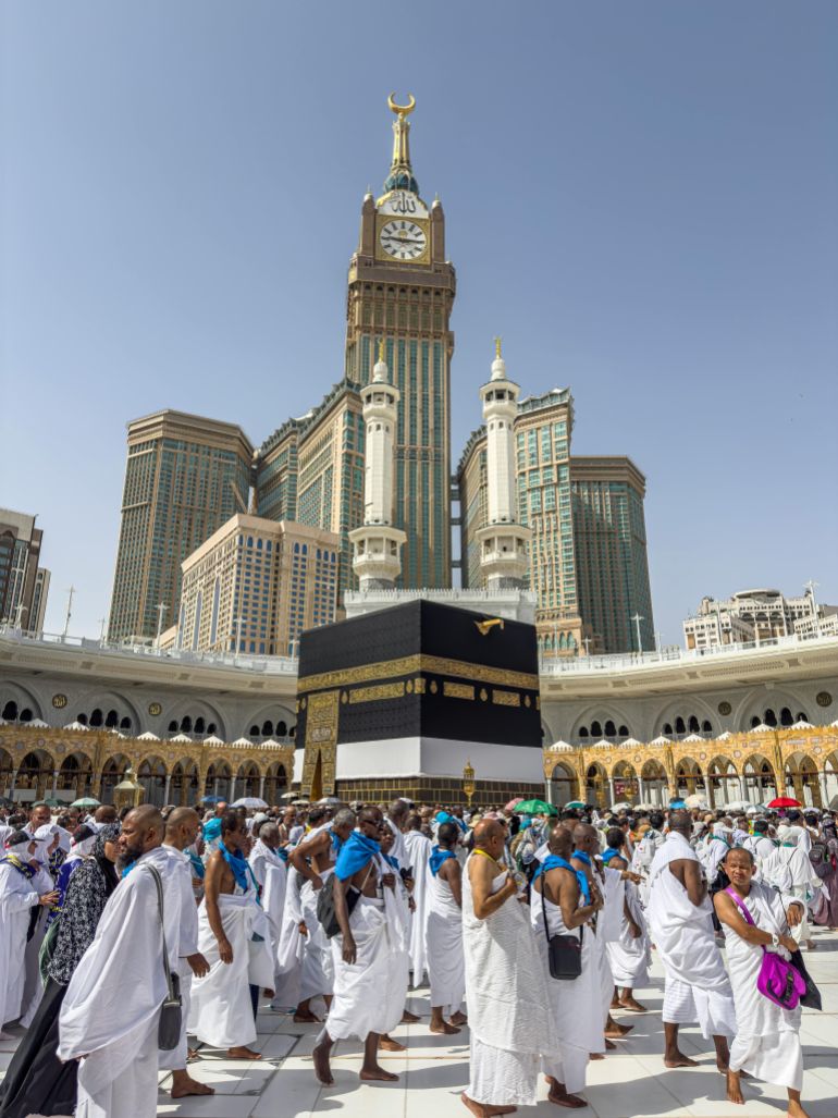 hajjj-1778756960 A large group of pilgrims gather at the Kaaba in Mecca, Saudi Arabia, during the Hajj pilgrimage under a clear blue sky.