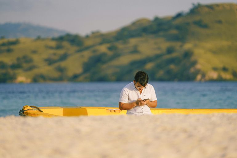 A man enjoys a peaceful moment with his smartphone by a yellow kayak on the beach in Badjo, Indonesia.