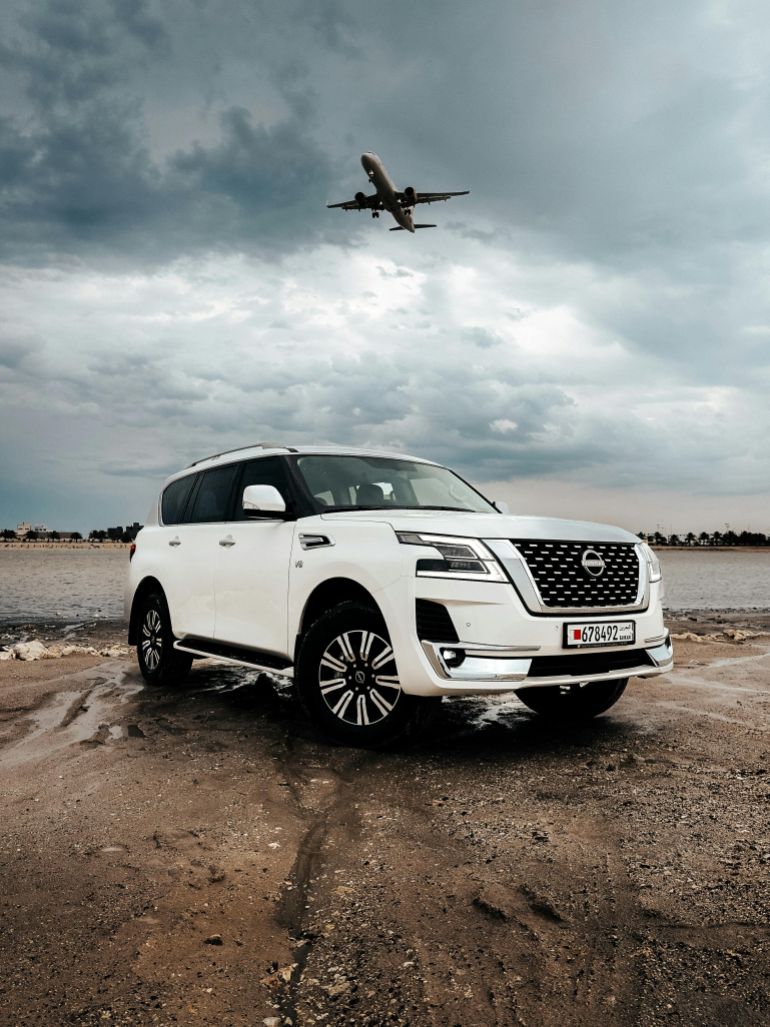 nysan-1779384838 White SUV parked on remote shoreline with airplane flying above on a cloudy day.