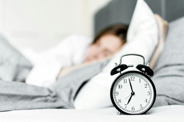 A retro alarm clock sits by a sleeping woman in a cozy bedroom setting.