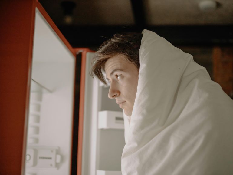 A man wrapped in a blanket checks an empty refrigerator at home, evoking a sense of isolation.