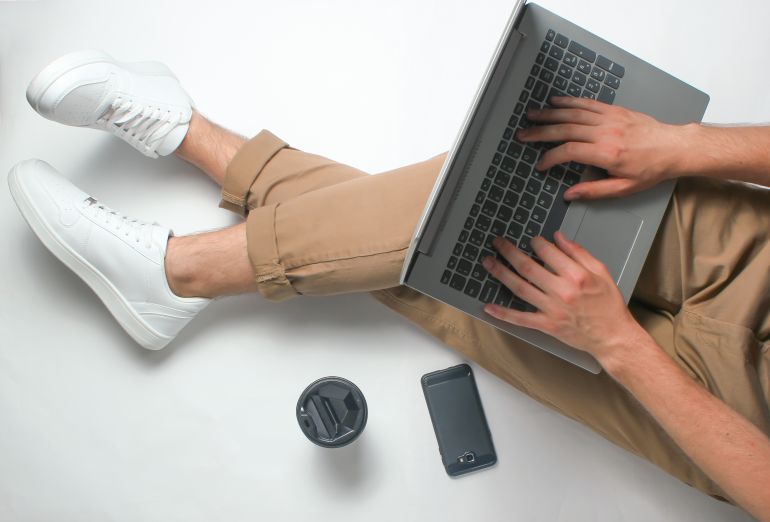Fragment of male legs in beige pants and white sneakers sitting on white. Man using modern laptop. Online worker, freelancing, work at home, man working. top view on white background