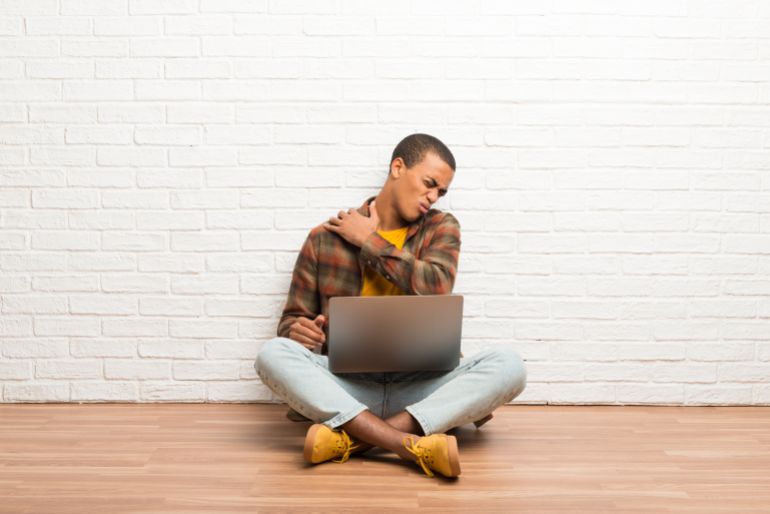 African american man sitting on the floor with his laptop suffering from pain in shoulder for having made an effort