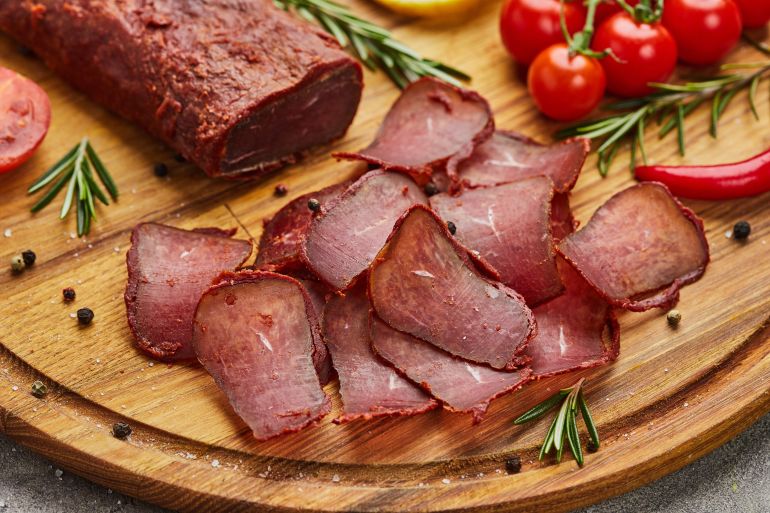 shutterstock_1715450113-1779265432 Armenian basturma or Pastirma on wooden cutting board decorated with spices, lettuce, lemon and tomatoes. Meat smoked jerky good as Beer snack