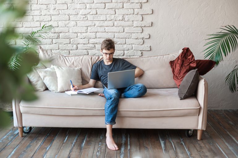 A teenage student boy writes notes sitting at home on the couch and listening to an online lesson. Distance education. Homeschooling. Self-education and self-discipline. Doing homework.