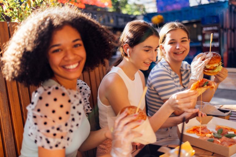 Cute smiling teenage girls are sitting in open air cafe and eating fast food. Cheerful happy female friends of different nationalities snacking on burgers and pizza outdoors and having fun together.