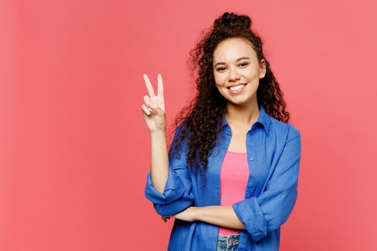 shutterstock_2356695649-1779100269 Young smiling happy cheerful woman of African American ethnicity she wear blue shirt casual clothes showing victory sign look camera isolated on plain pastel pink background studio. Lifestyle concept
