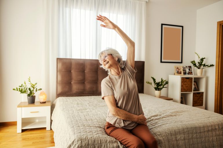 A happy senior woman sitting on a bed at home and doing senior fitness exercises.