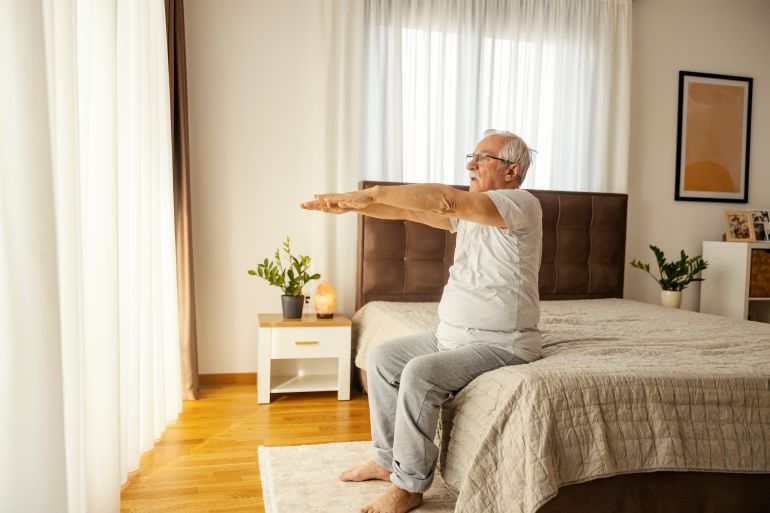 An active senior man is sitting on bed in a bedroom and doing morning workouts.