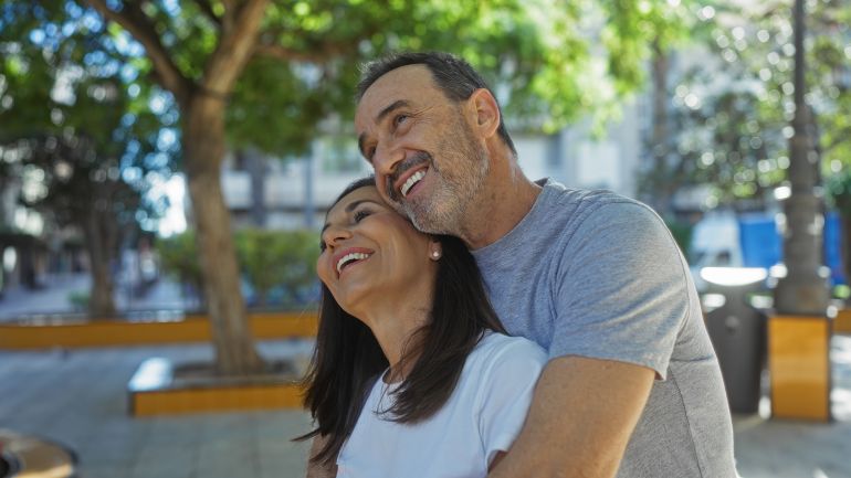 shutterstock_2601437267-1778411156 Middle aged hispanic couple embraces lovingly in a vibrant city park under lush trees, embodying joy and connection in the urban outdoors with a joyful smile.