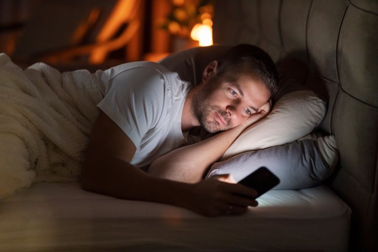 shutterstock_2602732133-1780228406 A sleepless man lying in dark bedroom with cellphone in hands and doom scrolling on social media.