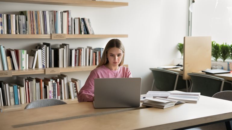 shutterstock_2608441685-1779627577 Elearning. Busy confident female student teenager work on notebook sitting at campus library desk check information received from paper book prepare coursework essay thesis conduct scientific research