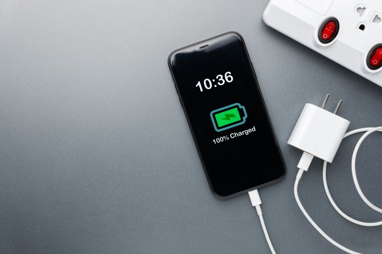 Mobile phone shows 100% charged battery connected to a USB cable with adapter unplugged from power strip. Concept highlights energy saving, smart power management
