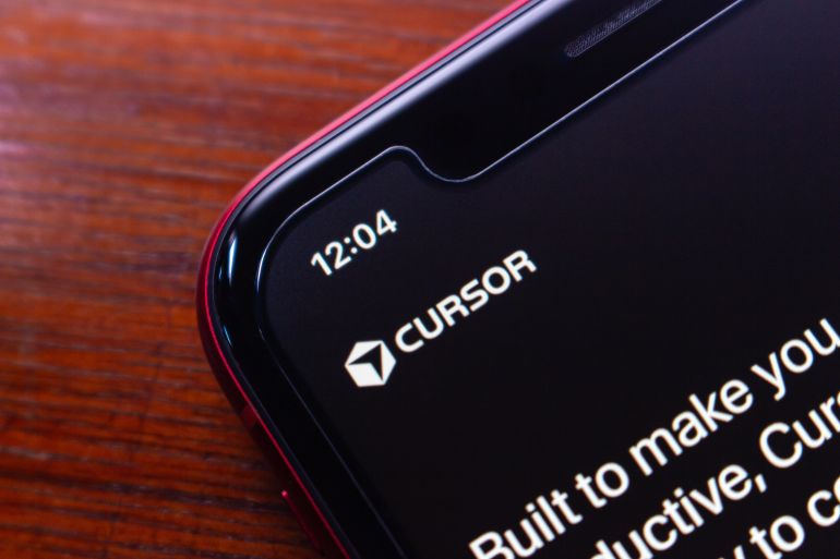 shutterstock_2710057979-1780220595 Quito, ECUADOR - Nov 25 2025 : Macro shot of a smartphone showing the Cursor app interface, emphasizing branding and modern user interface on a wooden surface.