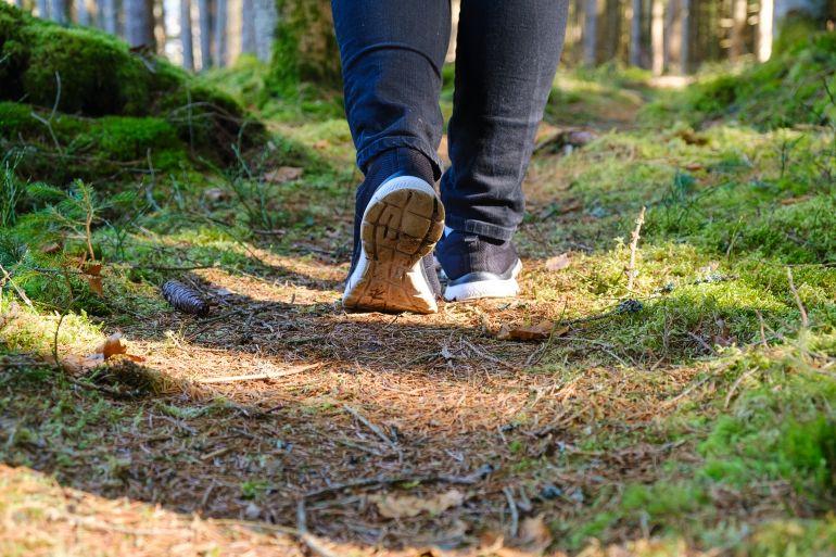 Low angle view of a person walking along a forest path. Focus on feet and trail with soft natural light and shallow depth of field. Depersonalized image representing outdoor activity, hiking and mindfulness in nature.
