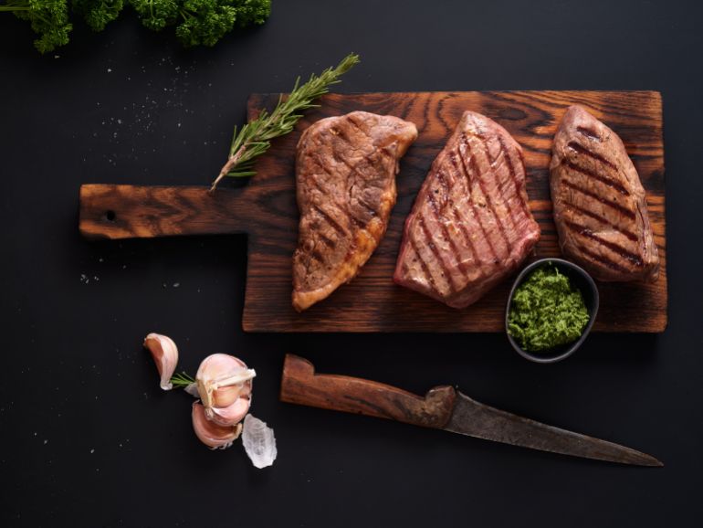 Grilled roast beef flank and rump steak are arranged on a wooden cutting board. Fresh herbs and garlic are nearby. A knife rests beside the board. This scene is set on a dark surface.