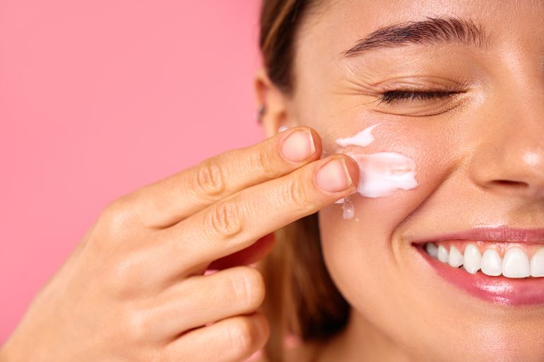 Caucasian woman with clear glowing skin using facial moisturizer or sunscreen isolated on pink background. Facial skin care. Spa treatments and cosmetology.