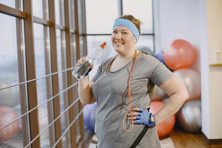 Smiling woman in gym with water bottle and jump rope, showing fitness motivation.