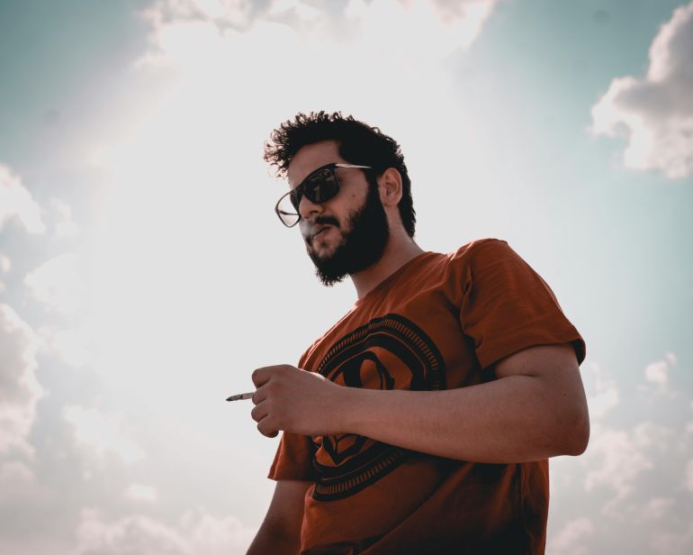 A bearded man wearing sunglasses smokes a cigarette under a bright sky.