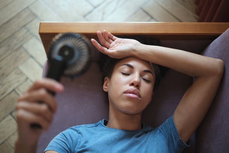 Close up of tired Exhausted African American woman lying in sofa using portable handheld mini fan suffering from summer heat, enjoying fresh air, cooling down at home