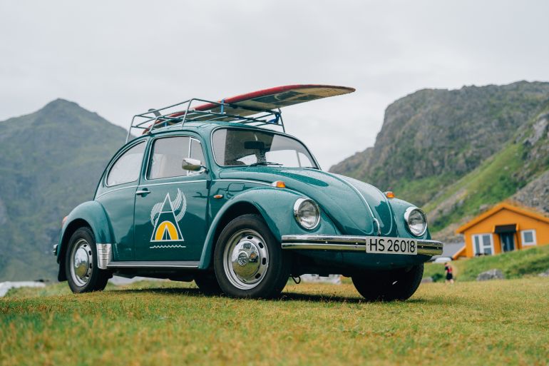 465sd-1780833359 Lofoten, Norway 17.6.2024: A vintage green Volkswagen Beetle with a surfboard on top, parked on a grassy field against a backdrop of rugged mountains.