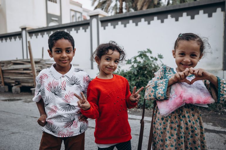5anas-1782046335 Three smiling children making peace signs in traditional clothing on a street in Nizwa, Oman.