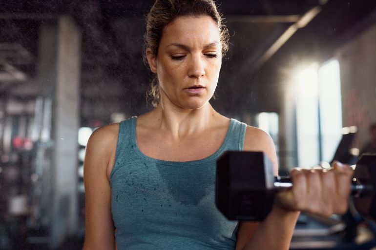 96-1782112294 Athletic woman making an effort while exercising strength with heavy dumbbell during sports training in a gym.