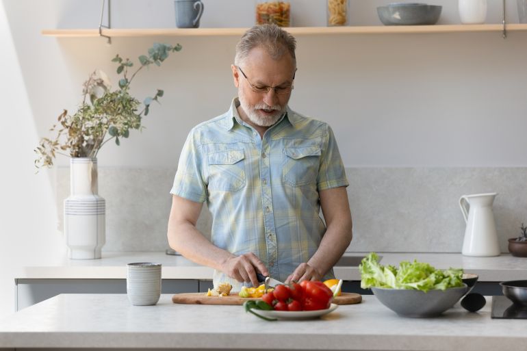 Senior grey haired vegetarian man cooking lunch in kitchen, cutting fresh vegetables at table, preparing organic meal, keeping healthy diet, caring of clean nutrition. Culinary, eating at home conceptالمصدر:أدوبي ستوك