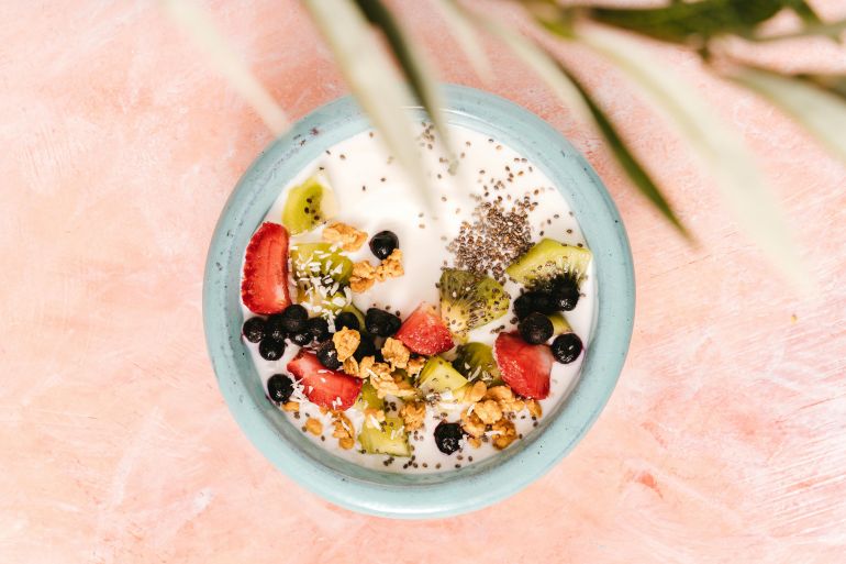 alshya-nzam-shy-1780754115 Top view of a healthy breakfast bowl with yogurt, fruits, and seeds on a pink background.