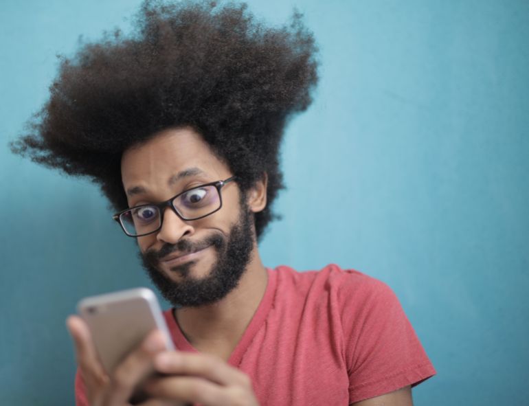 dhsht-alhatf-1781714517 A surprised man with afro hair and glasses looks at his smartphone against a blue background.