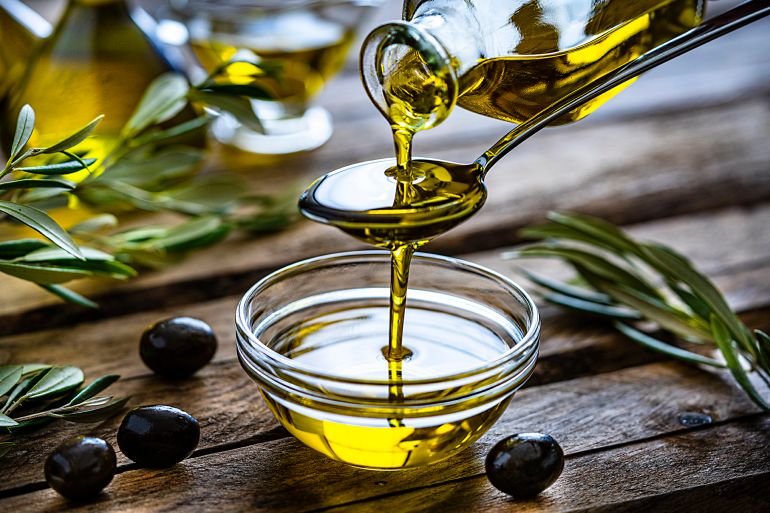 gettyimages-1206682746-1735831812 Pouring extra virgin olive oil from a spoon to a glass container. Olive branches comes from the left and right. Some olive oil bottles are out of focus at background. Black olives are on the table and complete the composition. The composition is on a rustic wooden kitchen table. Predominant colors are gold, green and brown. High resolution 42Mp studio digital capture taken with Sony A7rII and Sony FE 90mm f2.8 macro G OSS lens